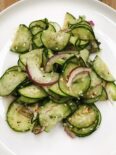 A white plate with vibrant green thin sliced cucumbers, and red onions, that have been tossed with a light vinaigrette and topped with sesame seeds.