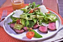 A jar of Lemon Vinaigrette dressing sits next to a plate with slices of seared flank steak, topped with romaine lettuce, shaved parmesan cheese and mixed marinated vegetables, including red onion, tomatoes, cucumbers, and snap peas.