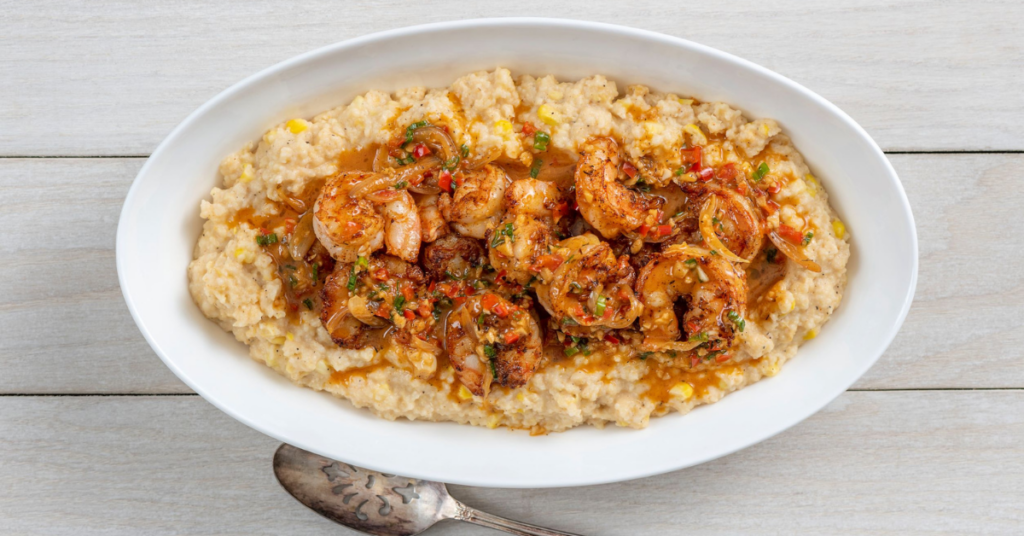 A white oval dish with shrimp cooked in spices, served on a bed of creamy grits, placed on a light wooden surface.